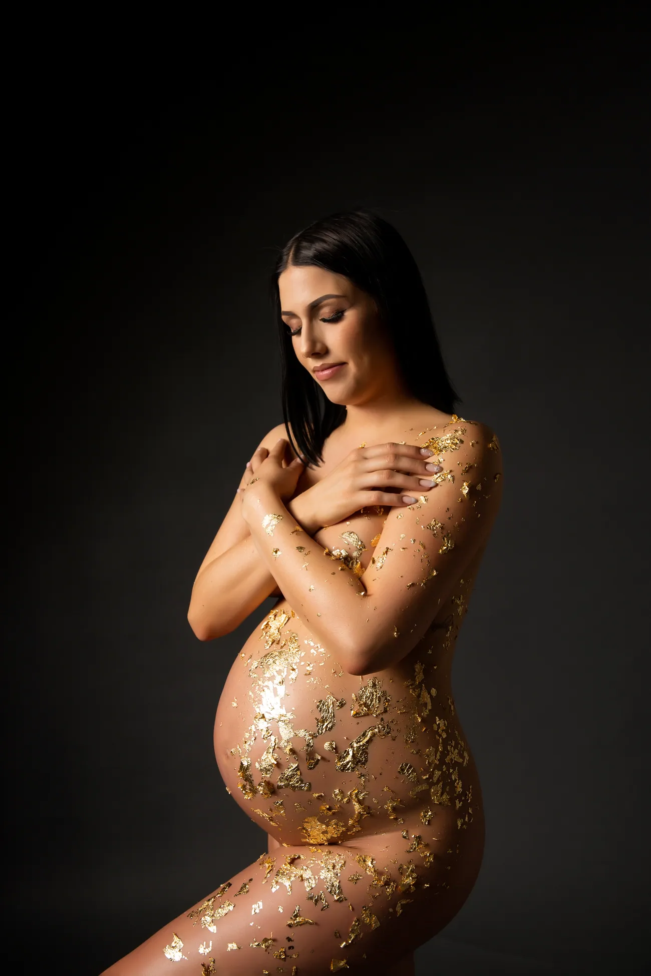 Babybauch-Portrait mit goldener Bemalung vor dunklem Hintergrund.