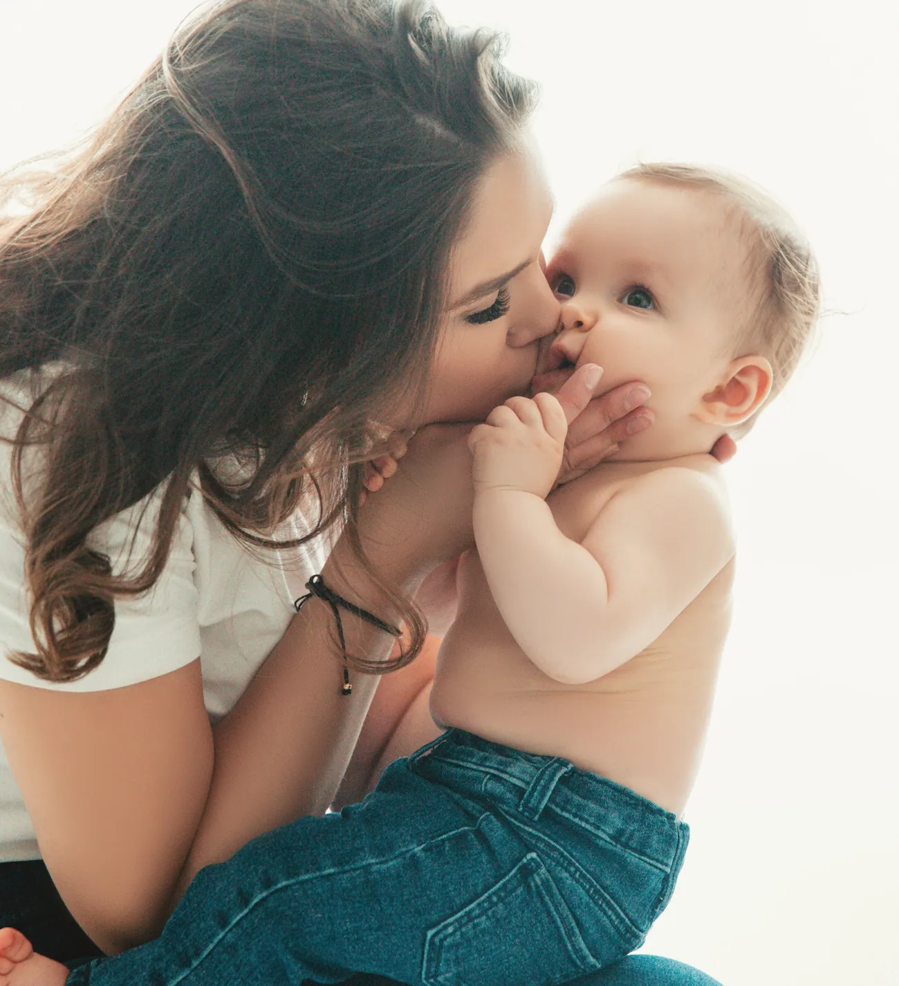 Mutter küsst lachend ihr Baby, im Gegenlicht, natürlicher Moment.