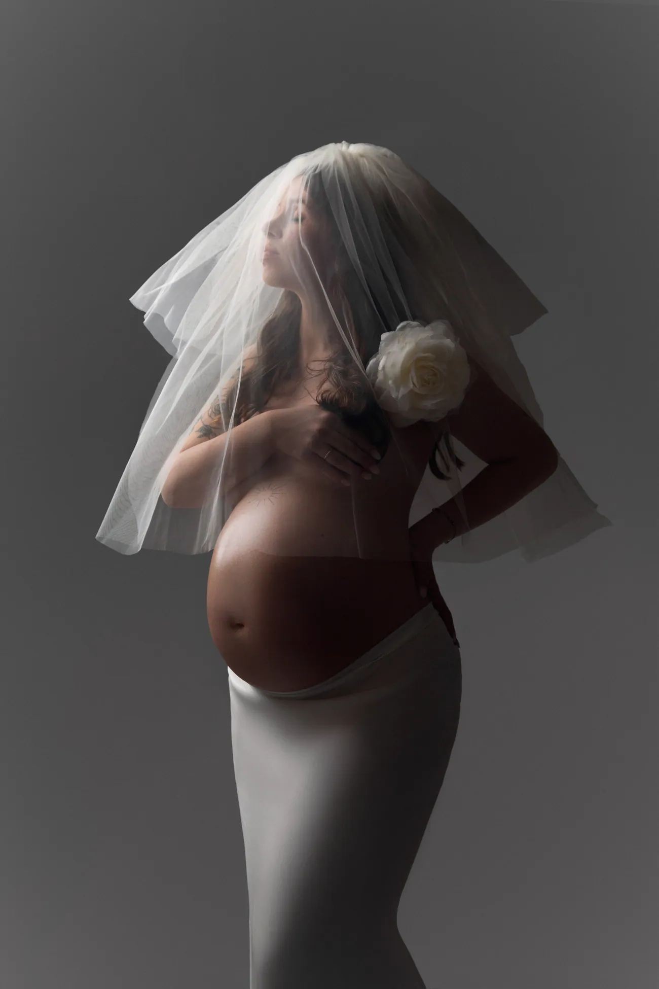 Babybauch-Portrait mit zartem Schleier vor grauem Hintergrund.
