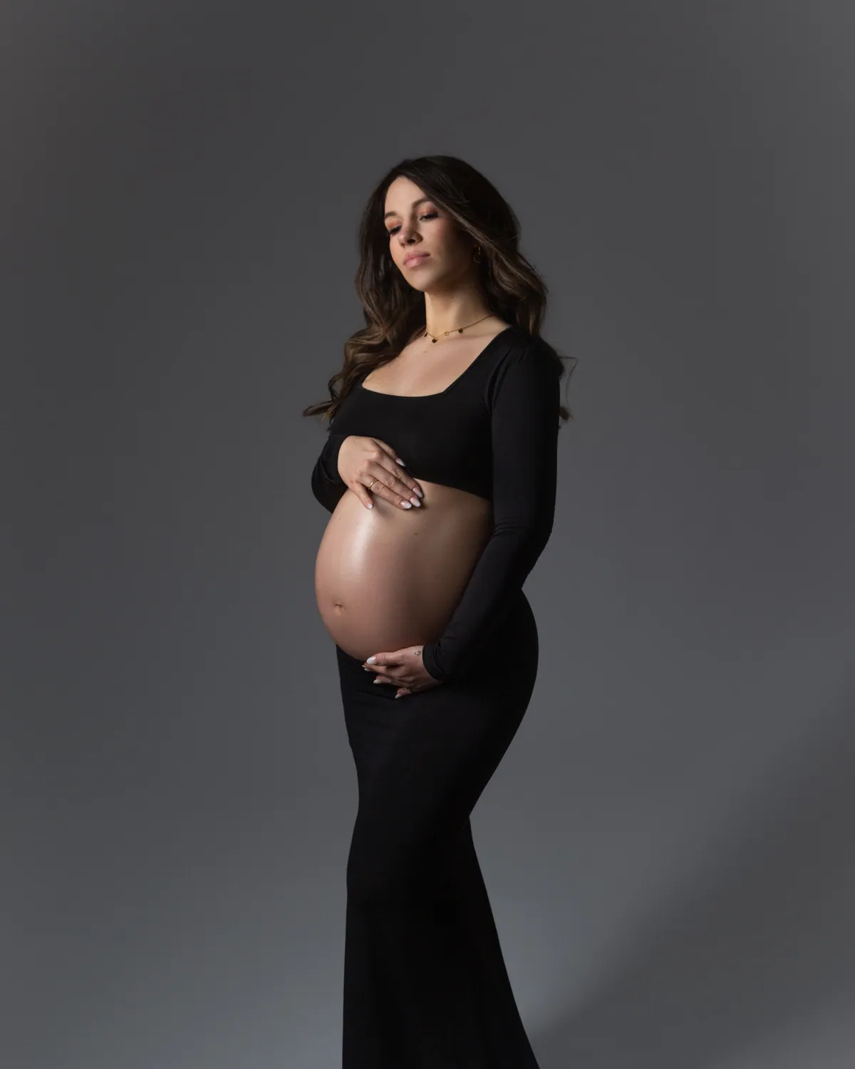 Schwangere Frau in einem fließenden schwarzen Kleid, elegantes Babybauch-Portrait vor grauem Studiohintergrund.